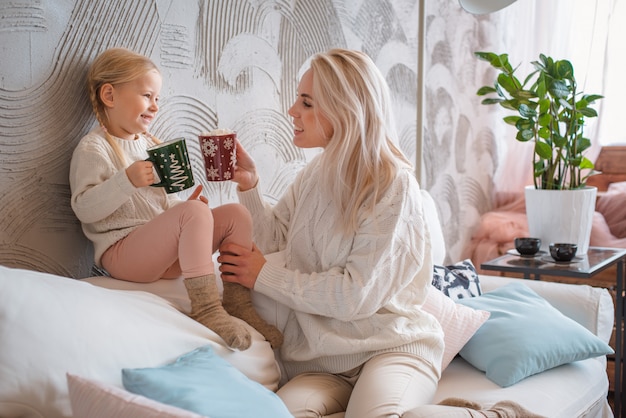 Young beautiful blonde woman hugs her little daughter on the couch with mugs of warm drink.