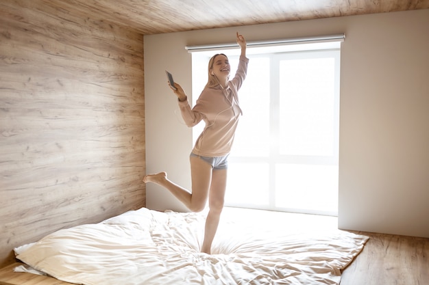 La giovane bella donna bionda salta sul letto questa mattina. il modello felice ascolta la musica tramite gli auricolari. telefono in mano. la luce del giorno esce dalla finestra.