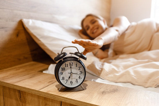 Photo young beautiful blonde woman lying in bed in morning. positive happy model reaching alarm clock with hand. . daylight. good mood.