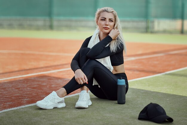 Young beautiful blonde woman sitting at tennis cort after doing fitness exercise. Girl rest after workout outdoors