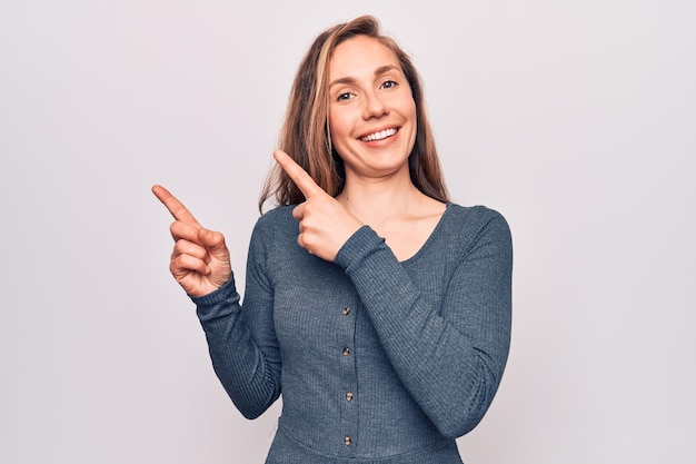 Young beautiful blonde woman standing over white isolated background smiling and looking at the camera pointing with two hands and fingers to the side