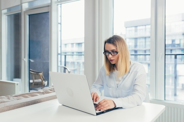 Young beautiful blonde woman typing on a laptop in the office tired looking at the camera