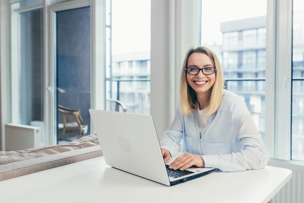 Young beautiful blonde woman typing on a laptop in the office tired looking at the camera