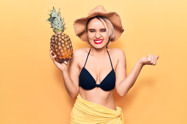 Young beautiful blonde woman wearing bikini and summer hat holding pineapple screaming proud celebrating victory and success very excited with raised arm