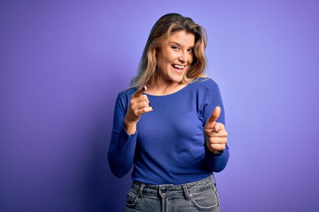 Young beautiful blonde woman wearing casual tshirt over isolated purple background pointing fingers to camera with happy and funny face good energy and vibes