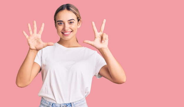 Young beautiful blonde woman wearing casual white tshirt showing and pointing up with fingers number eight while smiling confident and happy