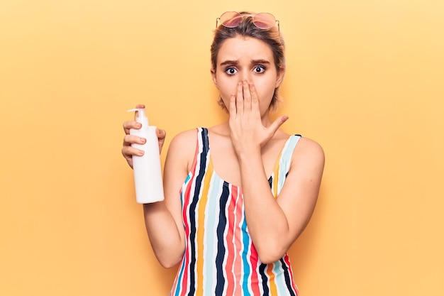 Young beautiful blonde woman wearing swimwear holding sunscreen lotion covering mouth with hand shocked and afraid for mistake surprised expression