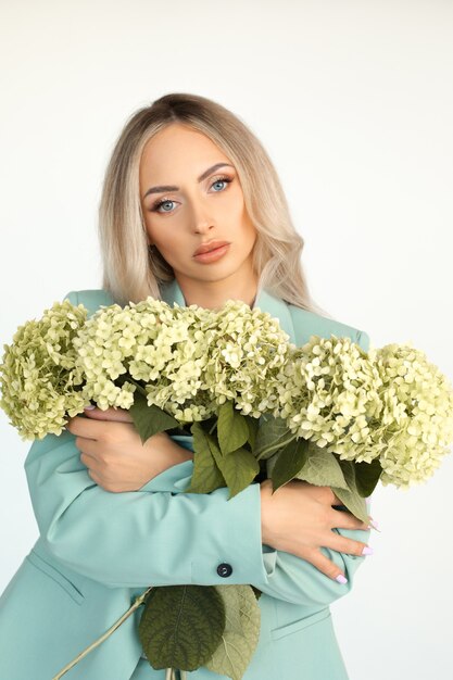 Young beautiful blonde woman with long hair in mint green pantsuit with armful of hydrangeas flowers in her arms. Portrait.