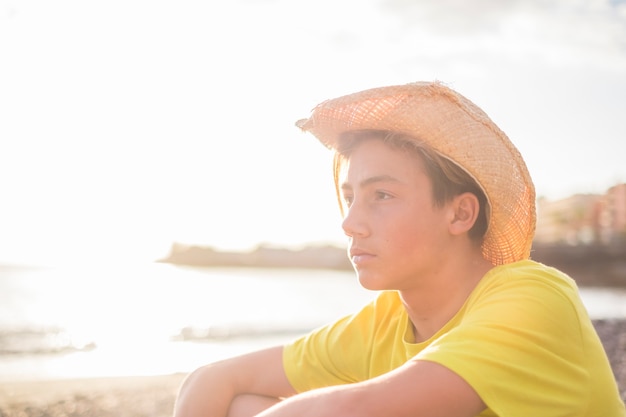 地平線を見ながら休暇でビーチに座っている15歳の若い美しい少年。海の波と夕日