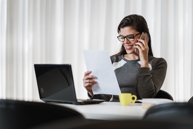 Young beautiful Brazilian woman using smartphone and working with laptop