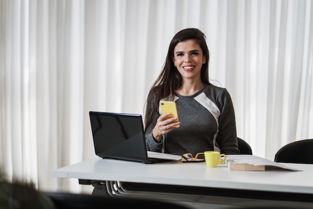 Young beautiful Brazilian woman using smartphone and working with laptop