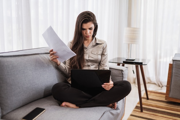 Young beautiful Brazilian woman working with laptop while sitting at sofa