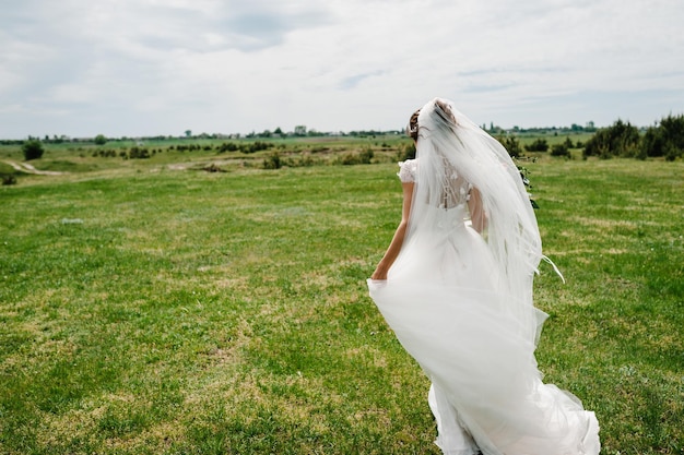 エレガントなドレスを着た若い美しい花嫁は、森の近くの野原に立ち、結婚式の後、自然の屋外で花束の花を持っています