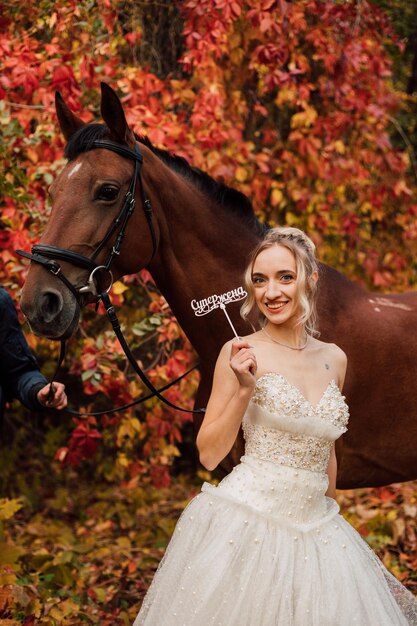 Young beautiful bride in a lush white dress happy walks with a horse in the autumn forest