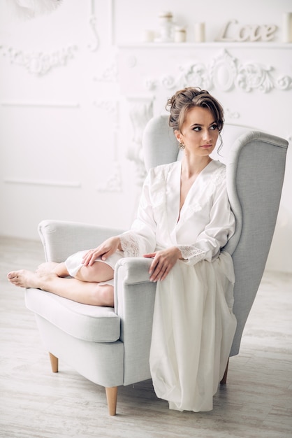 Young beautiful bride in a soft white robe sitting on a chair in a bright interior with candles on the background