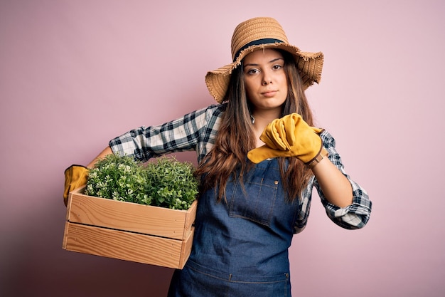 Young beautiful brunette farmer woman wearing apron and hat holding box with plants with angry face negative sign showing dislike with thumbs down rejection concept