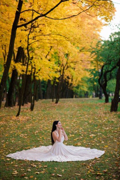 Young beautiful brunette female in a long white dress in autumn park