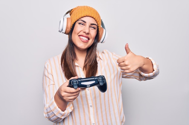 ジョイスティックとヘッドフォンを使ってビデオゲームをプレイする若い美しいブルネットのゲーマー女性は、幸せで肯定的な親指を上げ、優れた承認サインを行っています
