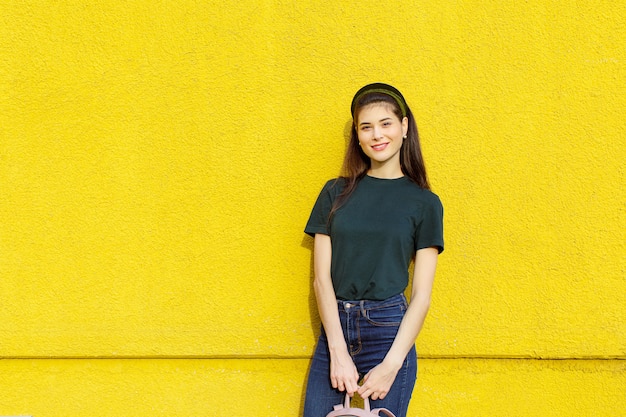 Young beautiful brunette girl in jeans a green T-shirt hair bands and a pink backpack posing on a yellow concrete wall