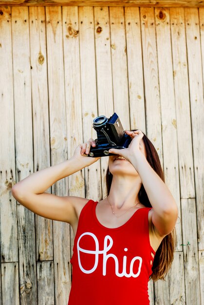 Young beautiful brunette girl in red shirt and jeans shorts posing with a camera on the rustic wooden background.