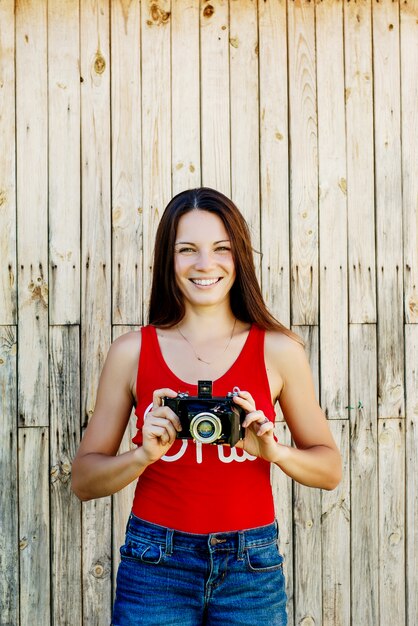 Young beautiful brunette girl in red shirt and jeans shorts posing with a camera on the rustic wooden background.