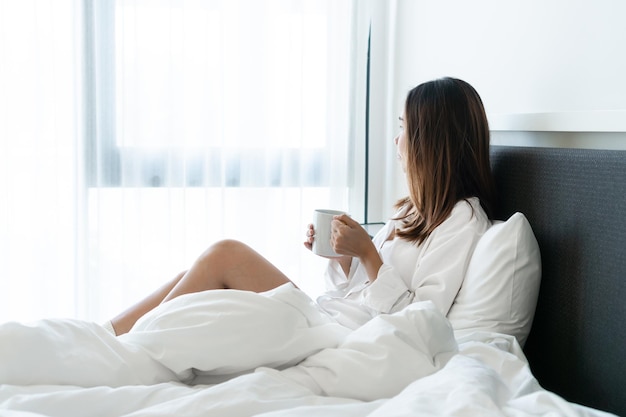 Young beautiful brunette hair woman in white shirt pajamas drinking coffee while sitting on the bed in the morning