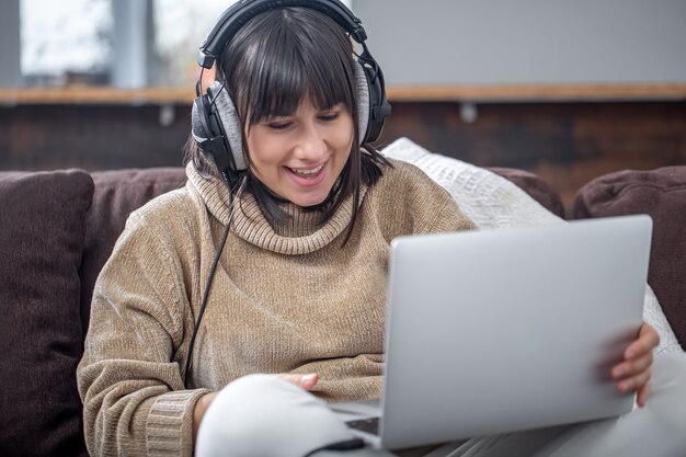 ヘッドフォンの若い美しいブルネットは、居心地の良いセーターに微笑んで、ノートパソコンの画面を見ています。オンライントレーニング、リモートワーク、ビデオコミュニケーションの概念。