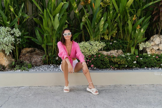 Young beautiful brunette in a pink shirt neck jewelry necklace trendy sunglasses sits on a background of tropical leaves laughs smiles