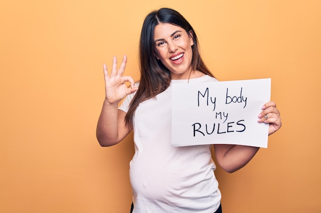 Young beautiful brunette pregnant woman holding banner with my body my rules message doing ok sign with fingers smiling friendly gesturing excellent symbol