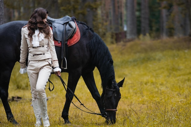 A young beautiful brunette rider next to a black horse in full ammunition portrait in a stable in a ...