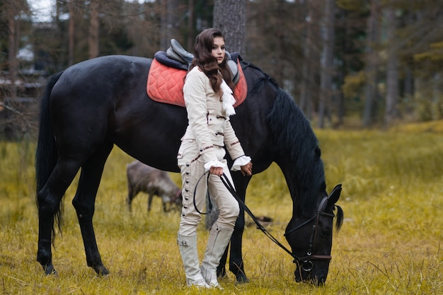 A young beautiful brunette rider next to a black mare in full ammunition,