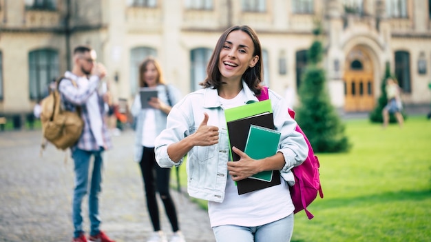 Young beautiful brunette student girl in denim clothes with backpack and books in hands on a group of her student friends