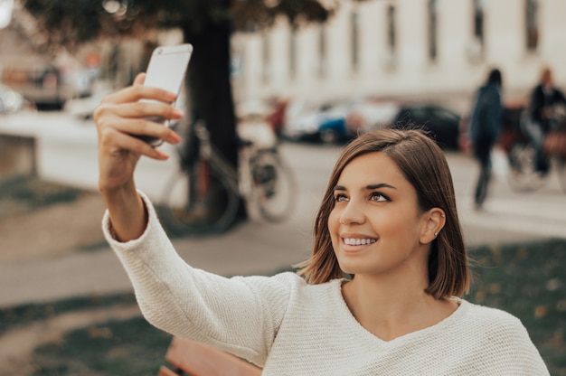 Young beautiful brunette taking pictures of herself on a cellphone in a city park.