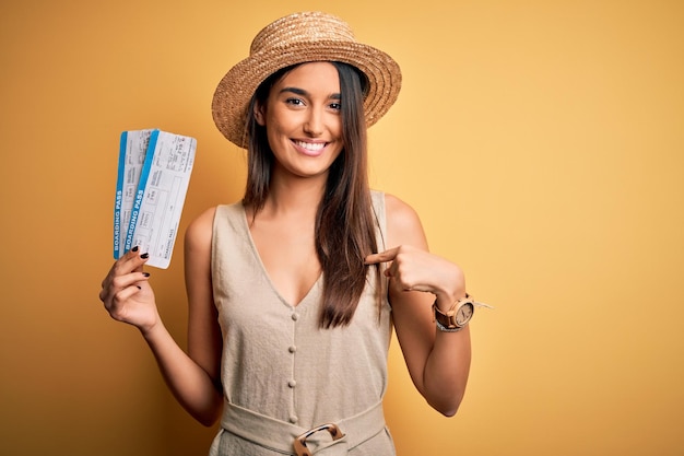 Young beautiful brunette tourist woman on vacation wearing hat holding boarding pass with surprise face pointing finger to himself