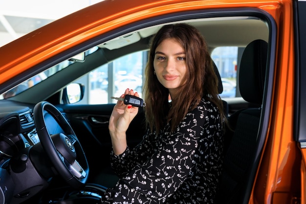 Young beautiful brunette while buying a new car A beautiful model in a dress sits behind the wheel of a car and shows the key to the camera