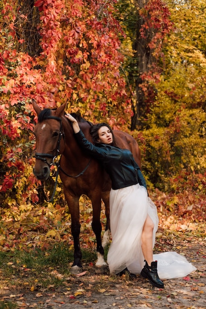 Young beautiful brunette in a white dress and a leather black jacket posing near a horse