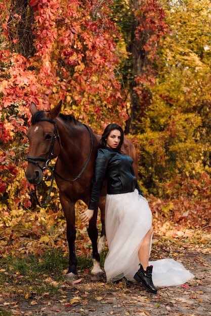 Young beautiful brunette in a white dress and a leather black jacket posing near a horse