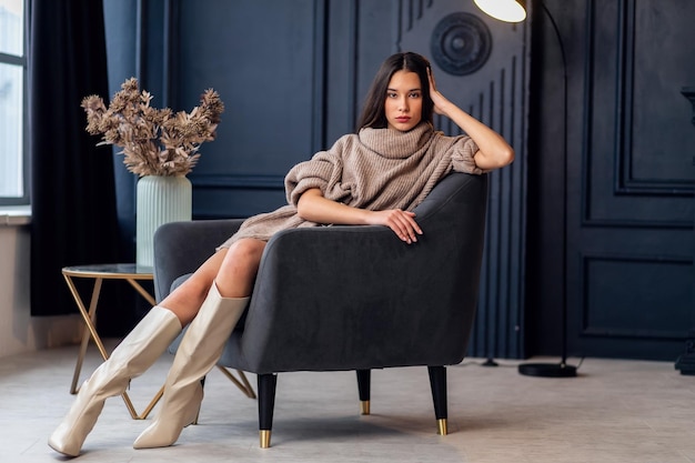 Young beautiful brunette woman in beige boots in a beautiful interior. Full length portrait. Young elegant woman wearing beige stylish look. Pretty girl with long brunette wavy hair. Fashionable lady