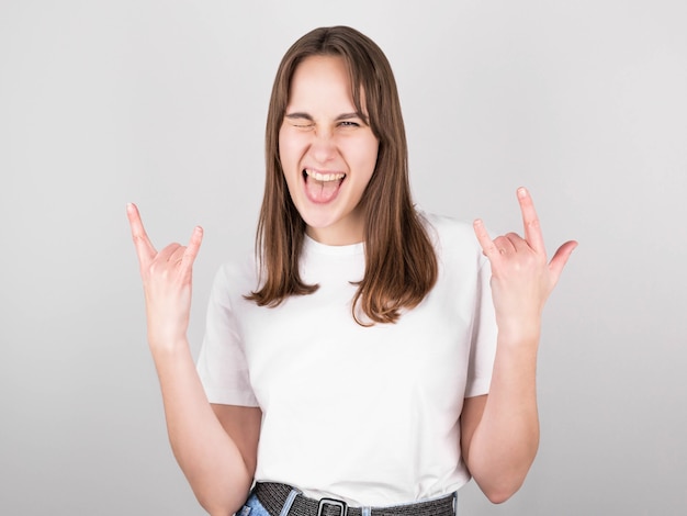 Young beautiful brunette woman in casual white t-shirt standing against isolated gray background, screaming with crazy facial expression