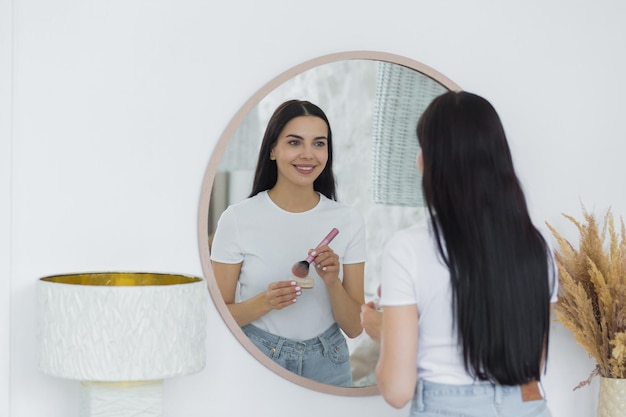 Una giovane bella donna bruna a casa si guarda allo specchio al mattino sorride applica il trucco