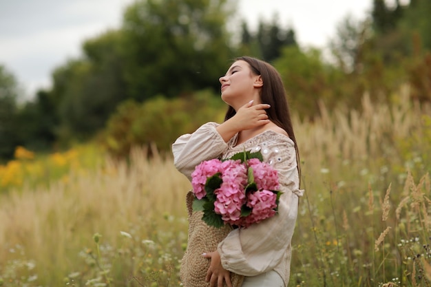 Young beautiful brunette woman is holding bouquet of pink flowers hydrangea in a straw bag,