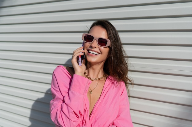 Молодая красивая брюнетка в розовой рубашке на шее, ювелирное ожерелье, модные солнцезащитные очки на фоне светлого гаражного забора, звонит по телефону, смеется, улыбается