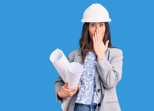 Young beautiful brunette woman wearing architect hardhat holding blueprint covering mouth with hand, shocked and afraid for mistake. surprised expression
