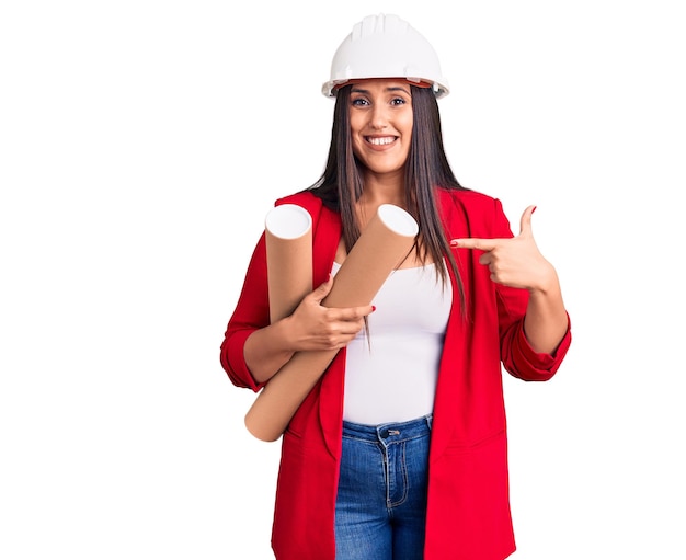 Young beautiful brunette woman wearing hardhat holding architect blueprint smiling happy pointing with hand and finger