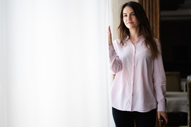 Young beautiful brunette woman at the window light background touches the curtain with her fingers