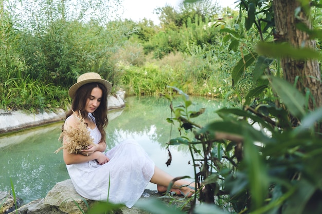 천연 수영장 옆에 맨발로 앉아 있는 젊은 아름다운 갈색 머리 여자. 하얀 선드레스를 입은 소녀와 꽃다발을 든 모자가 호수 옆의 돌 위에 앉아 있습니다.