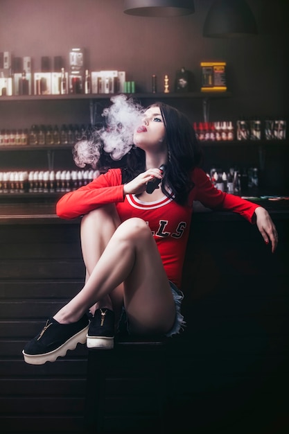 Young beautiful brunette woman with fashion makeup at the bar with a with vapor from electronic cigarette