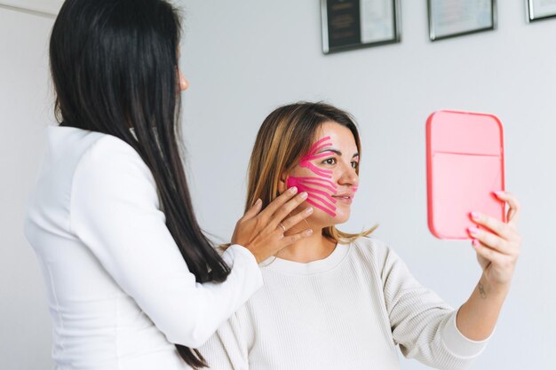 La giovane bella donna castana con il cosmetologo medico dei capelli lunghi fa il bendaggio facciale per il paziente in ufficio le giovani donne eseguono procedure antietà con l'aiuto del nastro kinesio