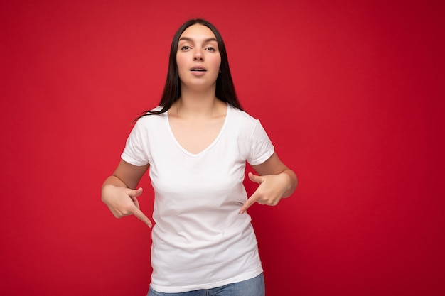 誠実な感情を持つ若い美しいブルネットの女性は、モックアップのためのカジュアルな白いTシャツを着て、空のスペースで指を指すコピースペースで背景の壁に分離されました。ポジティブなコンセプト。