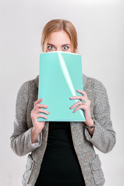 Young beautiful business woman blonde in black dress, jacket covers his face with a folder of papers and wonders on gray background